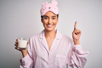 Young beautiful woman wearing pajama and sleep mask drinking glass of healthy milk surprised with...