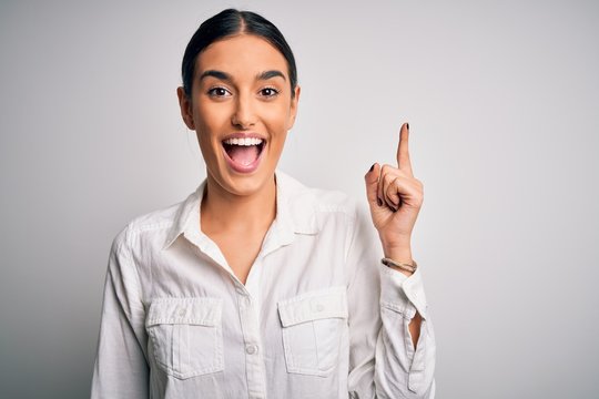 Young beautiful brunette woman wearing casual shirt over isolated white background pointing finger up with successful idea. Exited and happy. Number one.