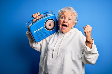 Senior beautiful woman holding vintage radio standing over isolated blue background screaming proud...