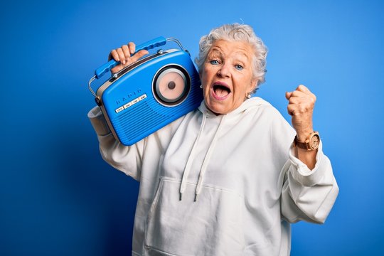 Senior beautiful woman holding vintage radio standing over isolated blue background screaming proud and celebrating victory and success very excited, cheering emotion
