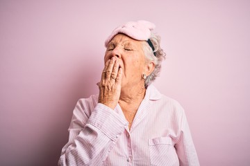 Senior beautiful woman wearing sleep mask and pajama over isolated pink background bored yawning...