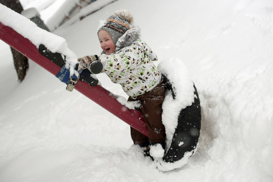 Small Happy Smiling Child With Red Cheeks And In Hat With Pompom Sitting On Swing And Laughing, Deep Snow Around