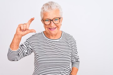 Senior grey-haired woman wearing striped navy t-shirt glasses over isolated white background smiling and confident gesturing with hand doing small size sign with fingers looking and the camera.