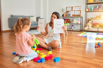 Beautiful psycologist and blond toddler girl sitting on the floor doing therapy using emoji emotions at consulting room