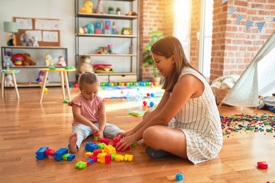 Beautiful teacher and blond toddler girl building tower using plastic blocks at kindergarten