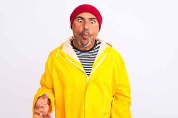 Middle age man wearing rain coat and woolen hat standing over isolated white background making fish face with lips, crazy and comical gesture. Funny expression.