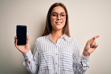 Young beautiful brunette woman holding smartphone showing screen over white background very happy pointing with hand and finger to the side