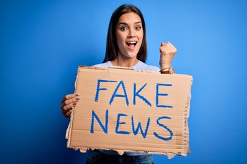 Young beautiful brunette woman holding banner with fake news message screaming proud and...