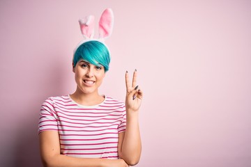 Young woman with fashion blue hair wearing easter rabbit ears over pink background smiling with happy face winking at the camera doing victory sign. Number two.