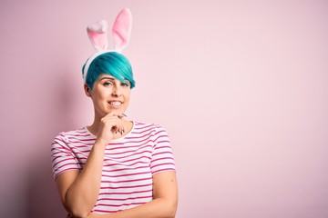 Obraz na płótnie Canvas Young woman with fashion blue hair wearing easter rabbit ears over pink background looking confident at the camera with smile with crossed arms and hand raised on chin. Thinking positive.