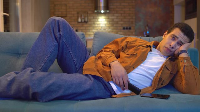 Tired guy watching tv home, yawning and falling asleep on couch late at night