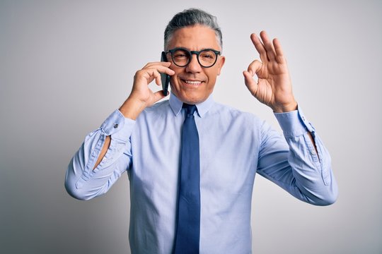 Middle age handsome grey-haired business man having conversation talking on the smartphone doing ok sign with fingers, excellent symbol