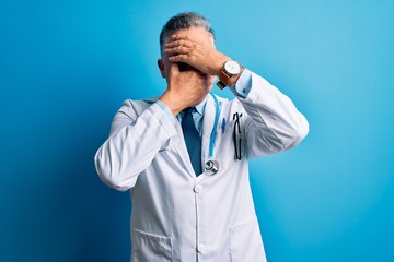 Middle age handsome grey-haired doctor man wearing coat and blue stethoscope Covering eyes and mouth with hands, surprised and shocked. Hiding emotion