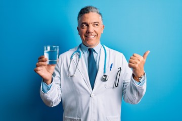 Middle age handsome grey-haired doctor man drinking glass of water over blue background pointing and showing with thumb up to the side with happy face smiling