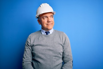 Middle age handsome grey-haired engineer man wearing safety helmet over blue background looking away to side with smile on face, natural expression. Laughing confident.