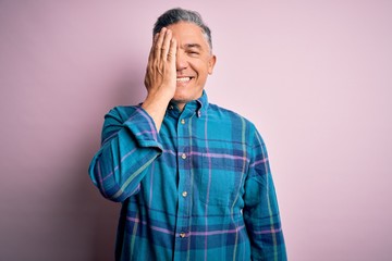 Middle age handsome grey-haired man wearing casual shirt over isolated pink background covering one eye with hand, confident smile on face and surprise emotion.