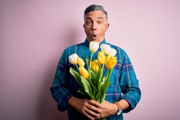 Middle age grey-haired man holding natural bouquet of yellow tulips over pink background scared in...