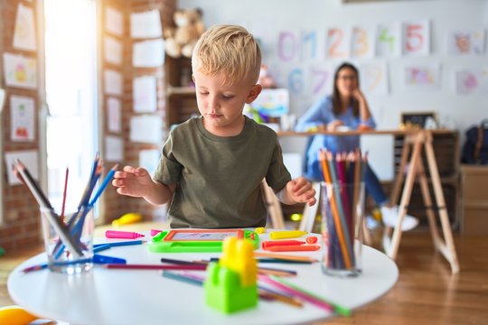 Young caucasian child playing at playschool with teacher. Mother and son at playroom drawing a draw with color pencils, young woman at the background sitting on desk.