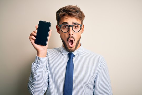 Young businessman holding smartphone standing over isolated white background scared in shock with a surprise face, afraid and excited with fear expression