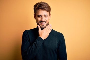 Young handsome man with beard wearing turtleneck sweater standing over yellow background looking confident at the camera smiling with crossed arms and hand raised on chin. Thinking positive.