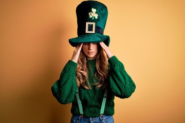 Beautiful brunette woman wearing green hat with clover celebrating saint patricks day with hand on head for pain in head because stress. Suffering migraine.