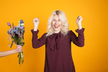 Excited blonde girl posing near tulip bouquet and showing yes gesture isolated on yellow background