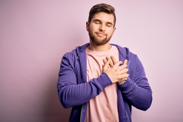 Young blond man with beard and blue eyes wearing purple sweatshirt over pink background smiling with hands on chest with closed eyes and grateful gesture on face. Health concept.