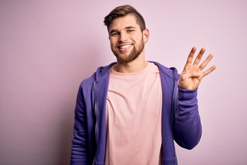 Young blond man with beard and blue eyes wearing purple sweatshirt over pink background showing and pointing up with fingers number four while smiling confident and happy.