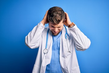 Young blond doctor man with beard and blue eyes wearing white coat and stethoscope suffering from headache desperate and stressed because pain and migraine. Hands on head.