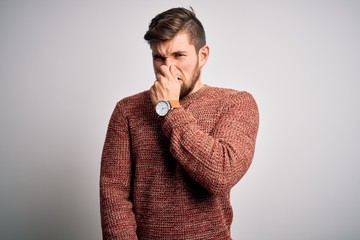 Young blond man with beard and blue eyes wearing casual sweater over white background smelling something stinky and disgusting, intolerable smell, holding breath with fingers on nose. Bad smell