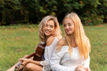 two young beautiful women friends outdoors playing a guitar and singing in park with sunset.