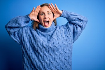 Young beautiful blonde woman wearing casual turtleneck sweater over blue background Smiling cheerful playing peek a boo with hands showing face. Surprised and exited