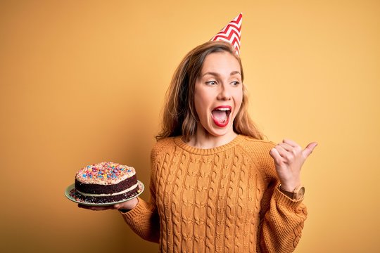 Young beautiful blonde woman holding birthday cake over isolated yellow background pointing and showing with thumb up to the side with happy face smiling