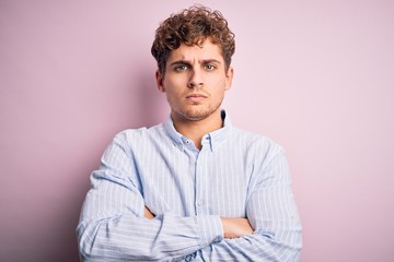 Young blond handsome man with curly hair wearing striped shirt over white background skeptic and nervous, disapproving expression on face with crossed arms. Negative person.