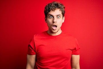 Young blond handsome man with curly hair wearing casual t-shirt over red background afraid and shocked with surprise and amazed expression, fear and excited face.