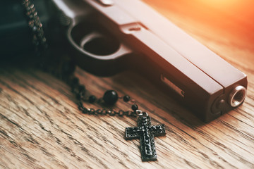 On a wooden table lies an airsoft black pistol and a black Gothic cross on a chain, illuminated by...