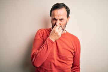 Middle age hoary man wearing casual orange sweater standing over isolated white background smelling something stinky and disgusting, intolerable smell, holding breath with fingers on nose. Bad smell