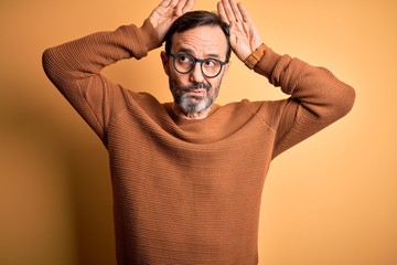 Middle age hoary man wearing brown sweater and glasses over isolated yellow background Doing bunny ears gesture with hands palms looking cynical and skeptical. Easter rabbit concept.