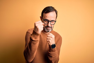 Middle age hoary man wearing brown sweater and glasses over isolated yellow background Ready to fight with fist defense gesture, angry and upset face, afraid of problem