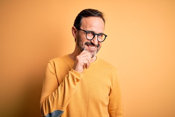 Middle age hoary man wearing casual sweater and glasses over isolated yellow background looking confident at the camera smiling with crossed arms and hand raised on chin. Thinking positive.