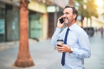 Middle age handsome businessman talking on the smartphone drinking take away coffee smiling