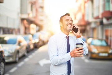 Middle age handsome businessman talking on the smartphone drinking take away coffee smiling