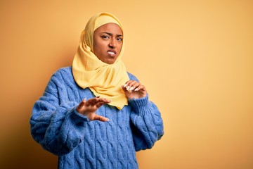 Obraz premium Young African American afro woman wearing muslim hijab over isolated yellow background disgusted expression, displeased and fearful doing disgust face because aversion reaction. With hands raised
