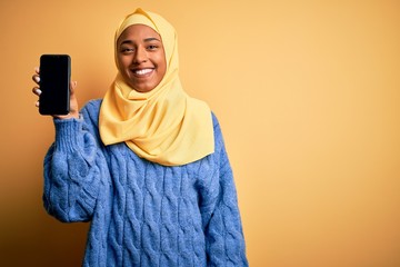 Young African American afro woman wearing muslim hijab showing smartphone with a happy face standing and smiling with a confident smile showing teeth