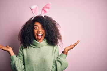 Young african american woman with afro hair wearing easter rabbit ears costume over pink background...