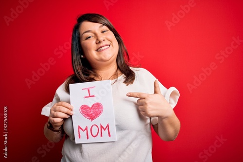 Beautiful brunette plus size woman holding love mom message celebrating mothers day very happy pointing with hand and finger