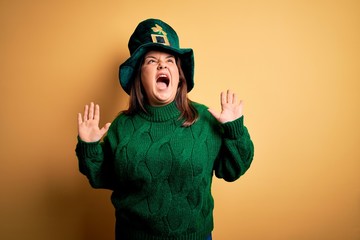 Young beautiful plus size woman wearing green hat with clover celebrating saint patricks day crazy and mad shouting and yelling with aggressive expression and arms raised. Frustration concept.