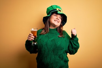 Young beautiful plus size woman wearing green hat celebrating st patricks day drinking beer pointing and showing with thumb up to the side with happy face smiling