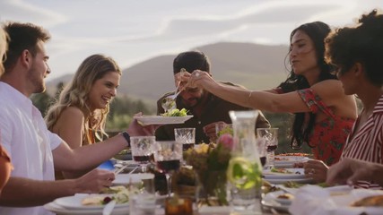 Woman serving food to friends having dinner at party table. Group of men and women having a dinner party outdoors. - Powered by Adobe