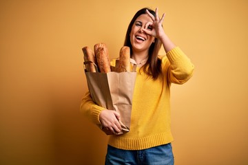 Young beautiful woman holding a bag of fresh healthy bread over yellow background doing ok gesture with hand smiling, eye looking through fingers with happy face.